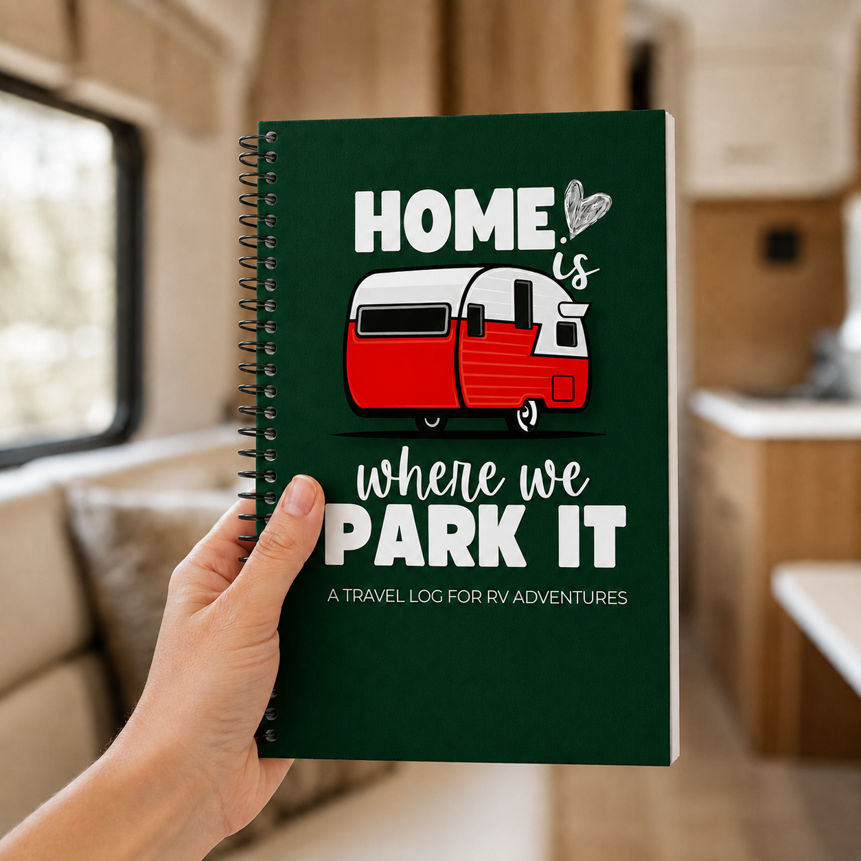 Hand holding a green Home Is Where We Park It RV travel journal inside a camper, showing its compact 6×9-inch size, spiral binding and vintage red travel-trailer cover.